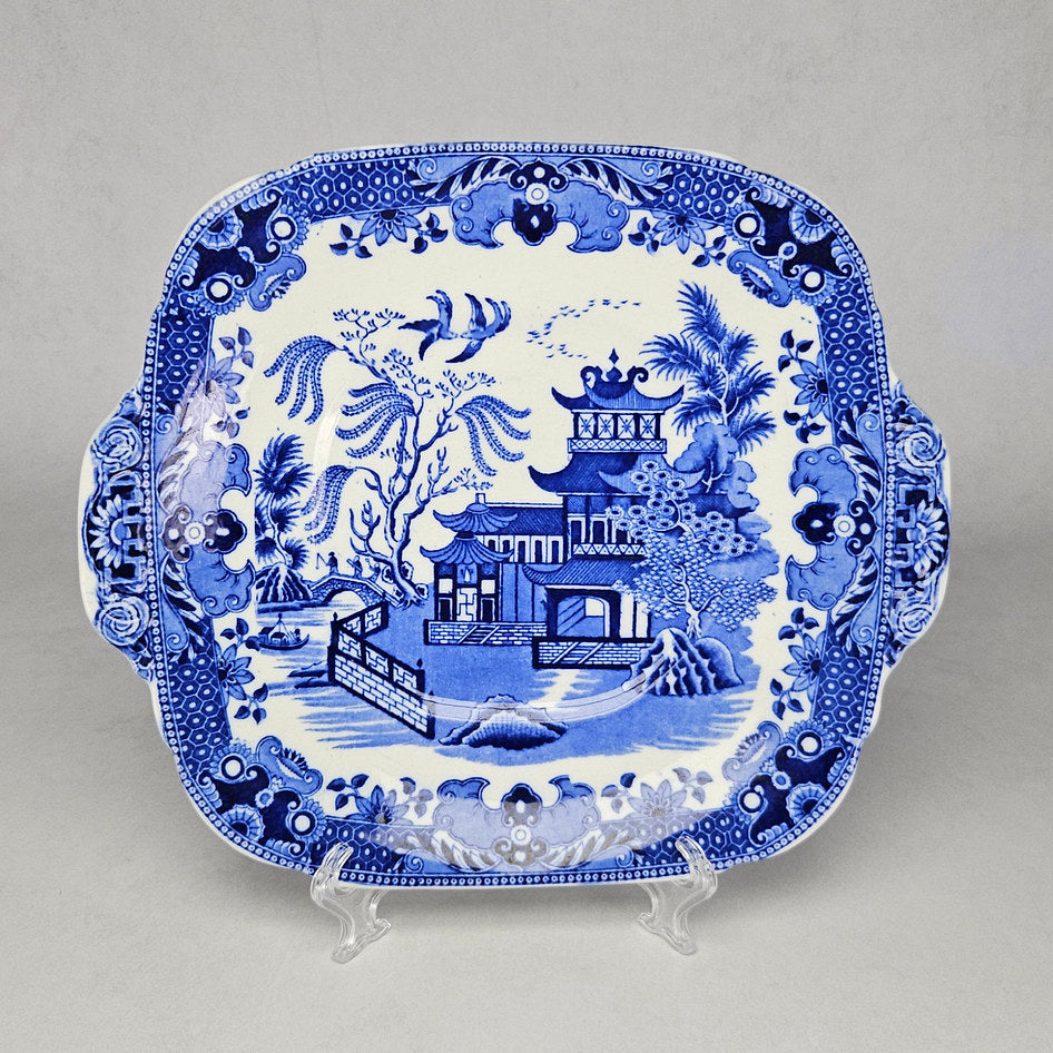 Burleigh Ware Willow blue and white rectangular handled serving tray displayed on a clear plate stand, showing the classic Blue Willow landscape scene with pagoda, bridge, birds and dense floral border